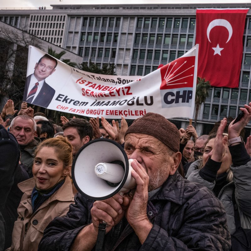 Eine Kundgebung in der Türkei. Ein Mann spricht in ein Megafon, er steht in einer Menschenmasse. Hinter ihm werden Transparente hochgehalten